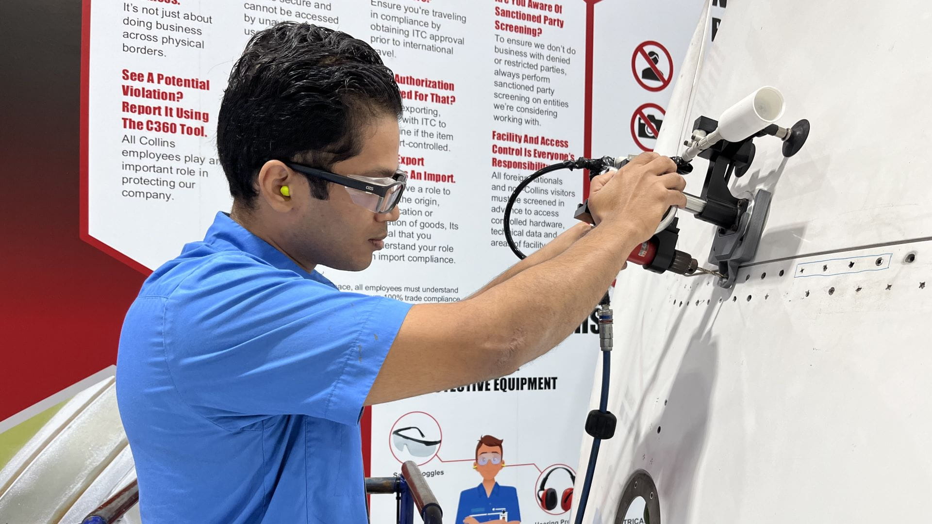 Young adult male wearing a blue short-sleeved collared shirt, yellow ear plugs and safety glasses operating a power drill.