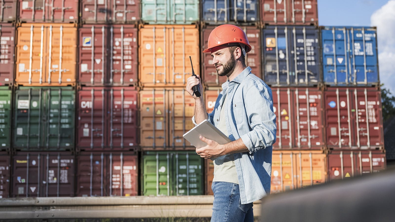 Middle-aged bearded adult male wearing an orange hard hat using a walky-talky with shipping containers in the background.