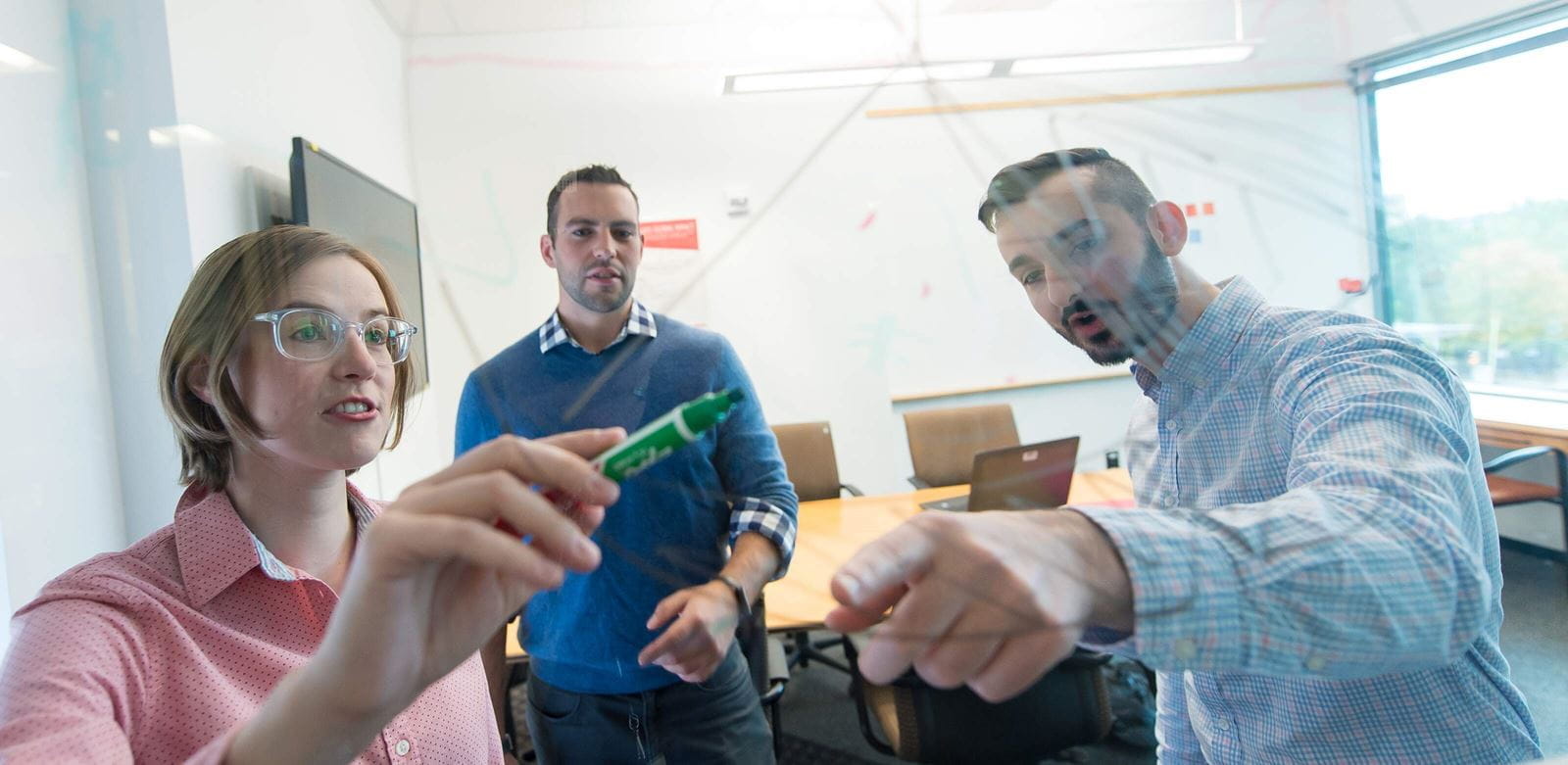 Two adult males and an adult female in a office setting using a whiteboard. 